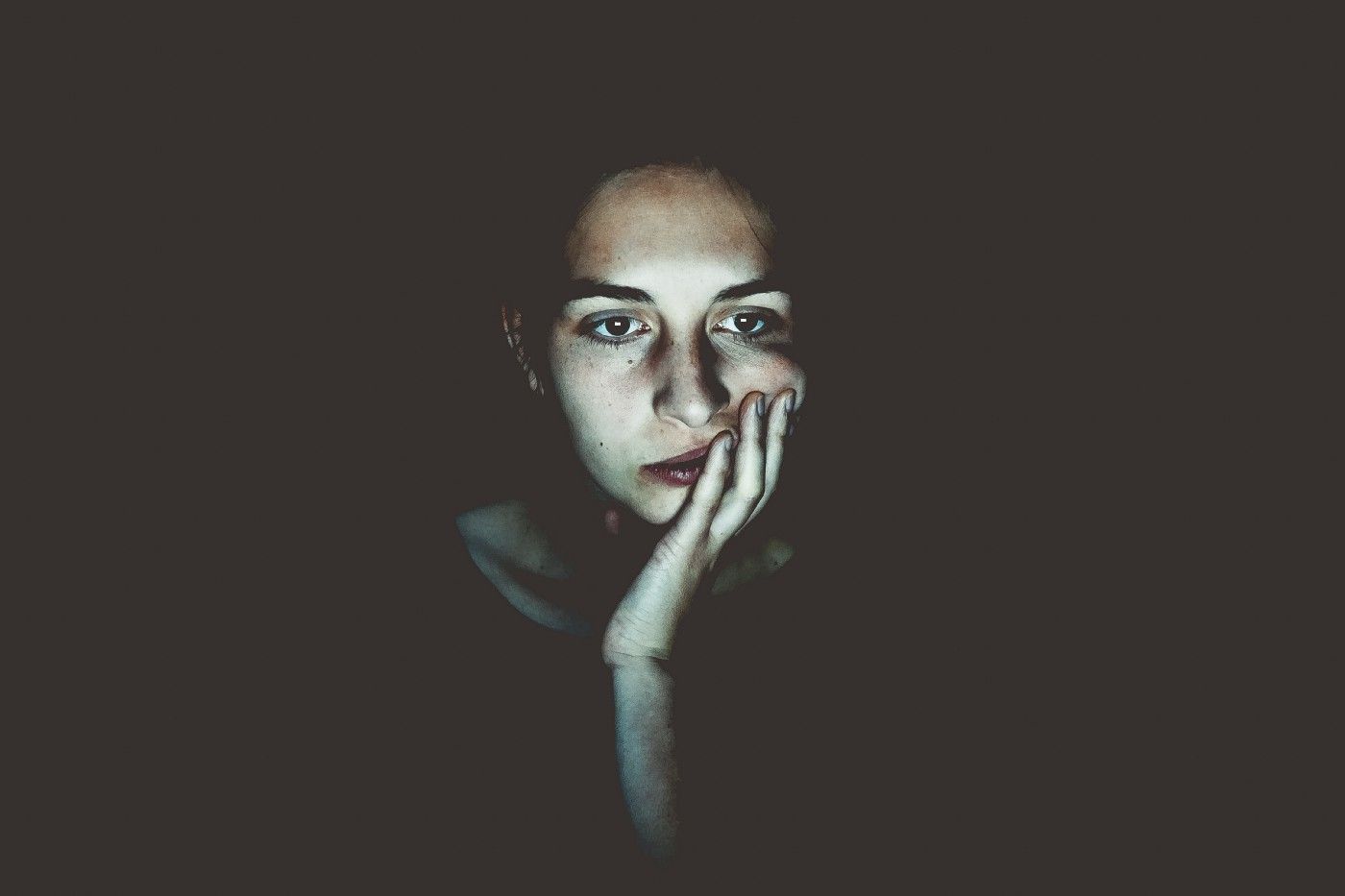 Woman in the dark looking down do a screen that reflects on her face. She looks tired.